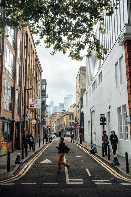 A narrow urban street scene in Shoreditch showing a variety of brick and modern white-painted buildings on either side. In the foreground, a person is walking across the road, while others are standing or sitting along the pavement. Street furniture such as bollards and double yellow lines mark the road edges. Several pedestrians are engaged in casual activities, with some waiting and others walking. Overhead, a large tree with green leaves extends into the frame, providing partial shade. The background features a view of high-rise buildings and a cloudy sky, indicating an urban environment with a mixture of older and contemporary architecture. The scene reflects an active area where private rubbish collection or on-site clearance services could be relevant, as suggested by the overall cityscape and the everyday street activity typical of central London neighborhoods like Shoreditch. Waste Disposal Shoreditch often services such busy streets for efficient rubbish removal and waste management solutions to support urban living and business needs.