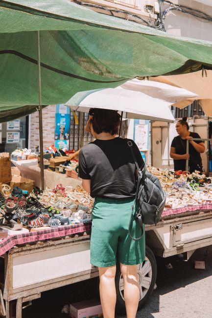 A woman with dark hair, dressed in a black t-shirt and bright green shorts, stands facing a street market stall under a green canopy, examining various small items, including watches and accessories displayed on a table covered with a pink checkered cloth. Behind her, a vendor with dark hair, wearing a black top, stands beside the stall, which is filled with an assortment of goods arranged in boxes and trays, some wrapped in plastic. The scene is set outdoors on a sunny day, with bright lighting and shadows cast on the pavement, and a background of urban storefronts and signs, reflecting a busy market environment typical of market traders handling diverse second-hand or vintage items, with Waste Disposal Shoreditch potentially involved in managing waste from such street markets, including discarded packaging and unsold stock.
