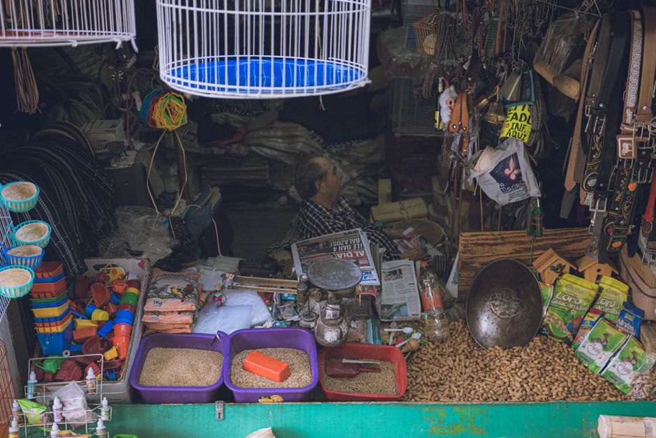 A market stall interior with a man seated behind a counter surrounded by various goods and clutter. The stall features stacks of packaged food items, containers filled with grains, and spices. To the right, there are hanging accessories such as belts, bags, and small ornaments. In the upper left, a glowing white circular light fixture illuminates the space, which has a dark background suggesting a compact shop or storage area. The foreground shows multiple plastic containers holding grains and powders, along with small colorful toys, and a large metal basin resting on a bed of dried food or small nuts. The scene portrays a busy, cluttered environment typical of local market stalls, with a focus on traditional trade and the collection or sale of various retail items, subtly connected to the theme of waste management through the assortment of packaging and materials present. Waste disposal and the handling of packaging materials are implied by the variety of items typically associated with local markets and independent collection practices.