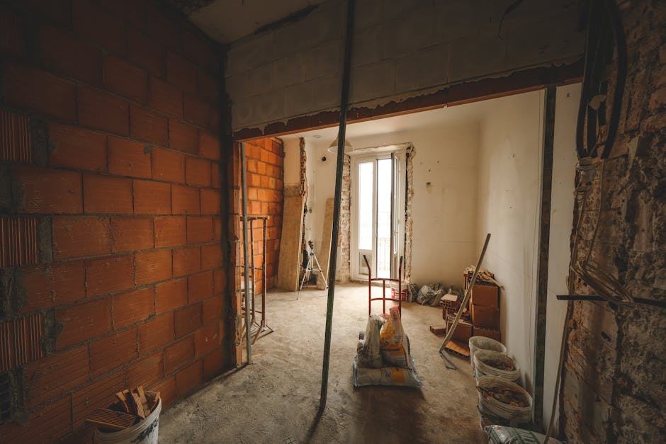 The interior of an under-construction or renovation space featuring exposed brick and plaster walls, with a large window at the far end allowing natural light to illuminate the room. The floor is unfinished concrete, scattered with construction materials and tools, including bags of cement, loose bricks, and wooden planks. In the foreground, two metal scaffolding frames support the ceiling opening, indicating ongoing structural work. To the right, an old rusted metal pulley system is mounted on the wall, and nearby, a small wooden shelf holds various materials, while a white plastic bucket sits on the floor. The overall scene suggests a workspace for building or renovation activities, with the presence of construction debris and the partial wall openings hinting at ongoing structural modifications, aligning with the context of private waste handling and on-site clearance services provided by Waste Disposal Shoreditch.